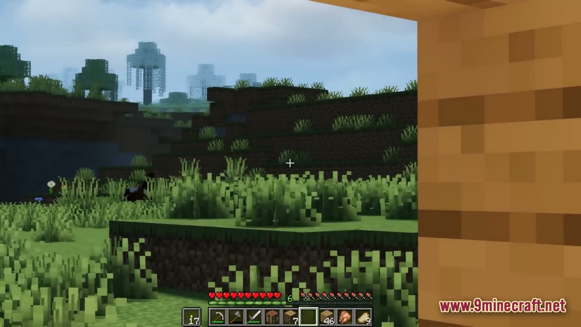A first-person view of a grassy minecraft landscape with distant structures and a player hotbar